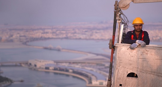 Un travailleur de la construction se tient debout dans un immeuble élevé, les gratte-ciels de Manama en arrière-plan.
