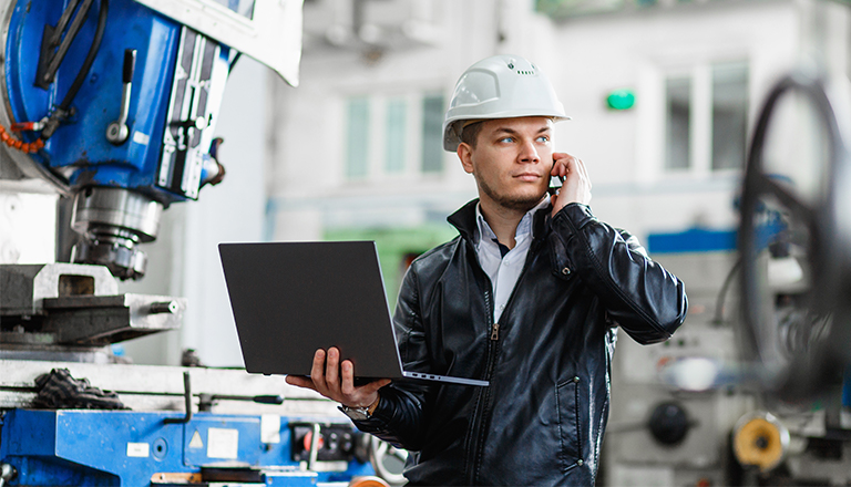 An engineer standing in front of an industrial machine with his laptop talks on the phone