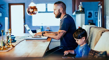 Man at a desk on his laptop with his young daughter sitting beside him.