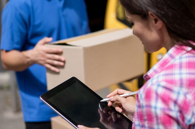 Two people stand in a warehouse discussing shipping.Waybill and shipping documentation are completed by a woman on tablet while man carries shipment.