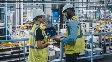 Male specialist and female car factory engineer in safety vests using laptop at vehicle production facility.