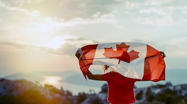 Person on mountaintop holding a Canadian flag over their head and looking out across landscape with sun shining in the distance.