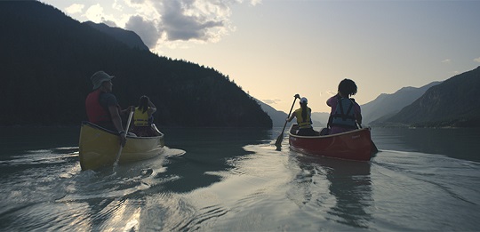 Two canoes, with two people in each, paddling in lake with mountain landscape.