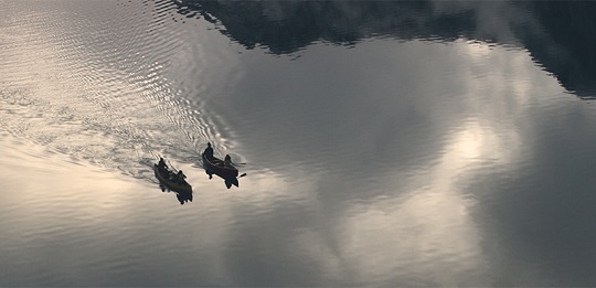 Two canoes, with two people in each, paddling side by side in open still water.