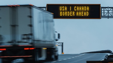 Transport truck passes under U.S.-Canada Border ahead sign on highway.