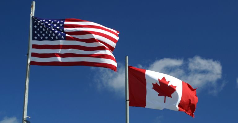 Les drapeaux des États-Unis et du Canada flottant au vent devant un ciel bleu et des nuages.
