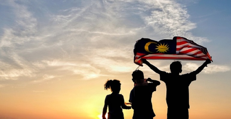 Silhouette de trois personnes, l’une tenant un drapeau malaisien, sur fond de coucher de soleil.