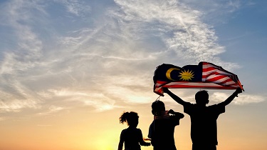 Silhouette de trois personnes, l’une tenant un drapeau malaisien, sur fond de coucher de soleil.
