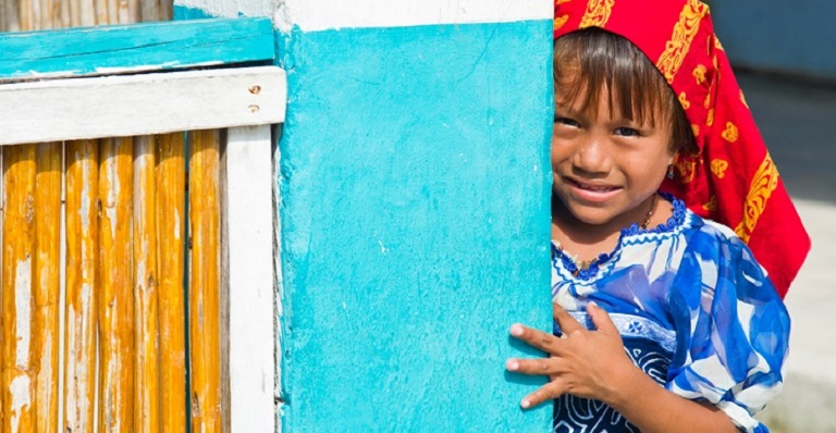 Un enfant portant une coiffe sourit à l’objectif, dissimulé derrière une porte.