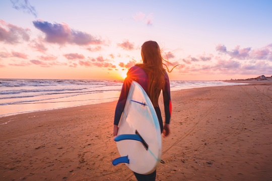 Surf girl with surfboard on a beach at sunset.