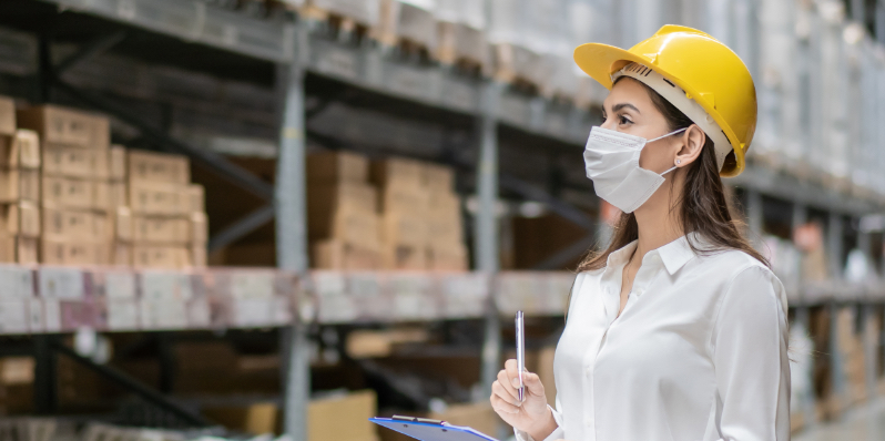 Essential worker in service and delivery industry wearing a face mask, walking through a warehouse taking inventory. 