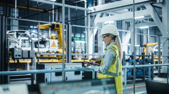 Une femme portant un casque, des lunettes et un gilet de sécurité utilisant un ordinateur portable, avec en arrière-plan un bras robotisé fabriquant un véhicule flou.