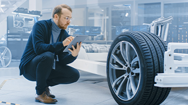 Un homme qui tient une tablette inspecte l’assemblage des roues et d’un châssis d’automobile dans une usine.