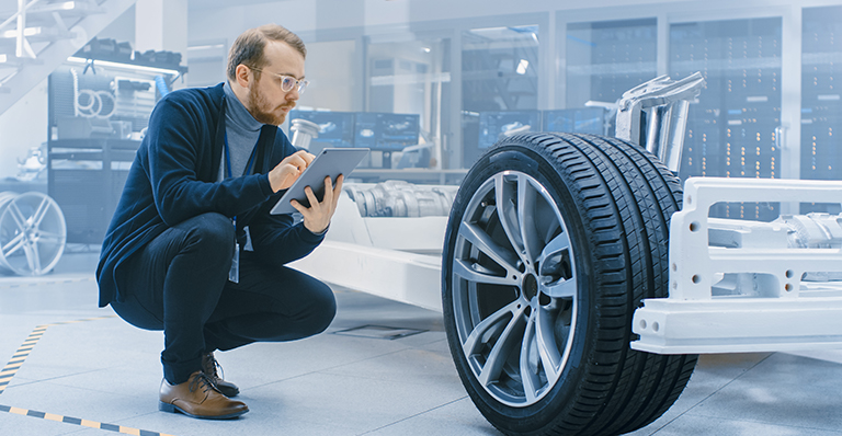 Man holding tablet inspects an automobile frame and wheel assembly in a factory.