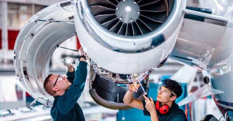 Two maintenance engineers inspect a jet engine inside an aircraft maintenance hangar.
