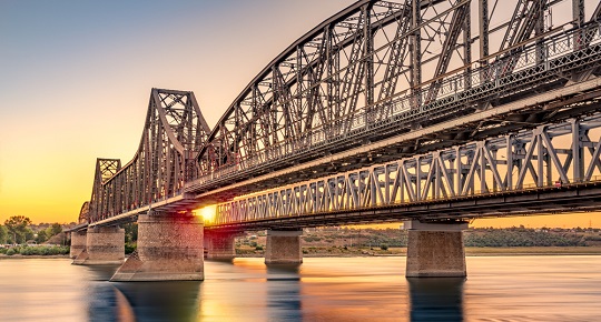 A long steel bridge stretches over a river into a busy city.