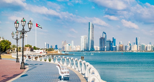 Un trottoir de bois s’étend le long d’un cours d’eau, sous un beau soleil et la ville de Dubaï en arrière-plan.