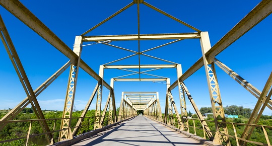 Un pont jaune en acier s’étend vers une zone densément boisée.