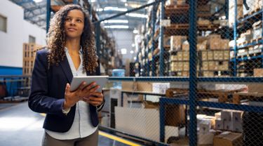A businesswoman in professional attire using a tablet in a warehouse with blurred shelved boxes behind her.