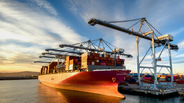 Aerial shot of a massive cargo ship loaded with containers docked in the Port of Long Beach, with the booms of ship-to-shore gantry cranes reaching out from the pier of the terminal.