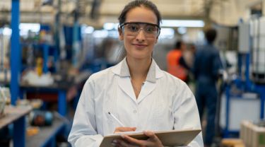 A smiling woman in a lab coat and safety glasses holding a clipboard and pen, standing in a manufacturing room.