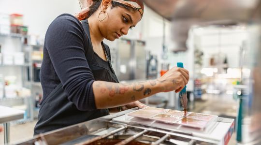 A woman chocolatier spreading melted chocolate into a bar mold inside a chocolate factory.