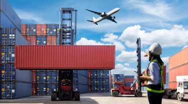 Un avion vole au-dessus de conteneurs et de camions, et une personne portant un gilet de sécurité se trouve au sol.