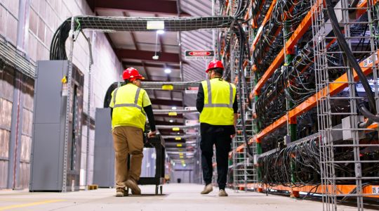 Two workers in safety helmets and high-visibility vests walk through an industrial data centre aisle lined with server racks and bundled cables.