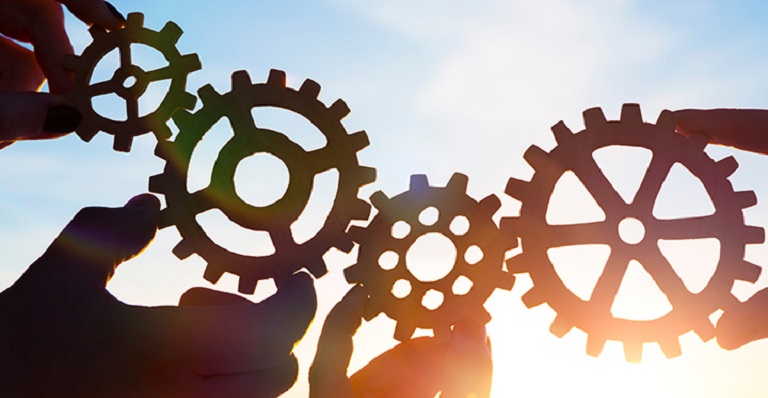Four hands, each holding a gear, work together to move forward against a sunlit sky.