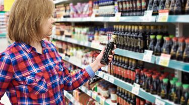 A woman holding a bottle of soy sauce in a grocery store aisle filled with various soy sauce bottles and condiments.