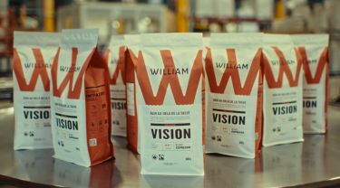 Packaged Café William espresso beans in focus with a factory background softly blurred.