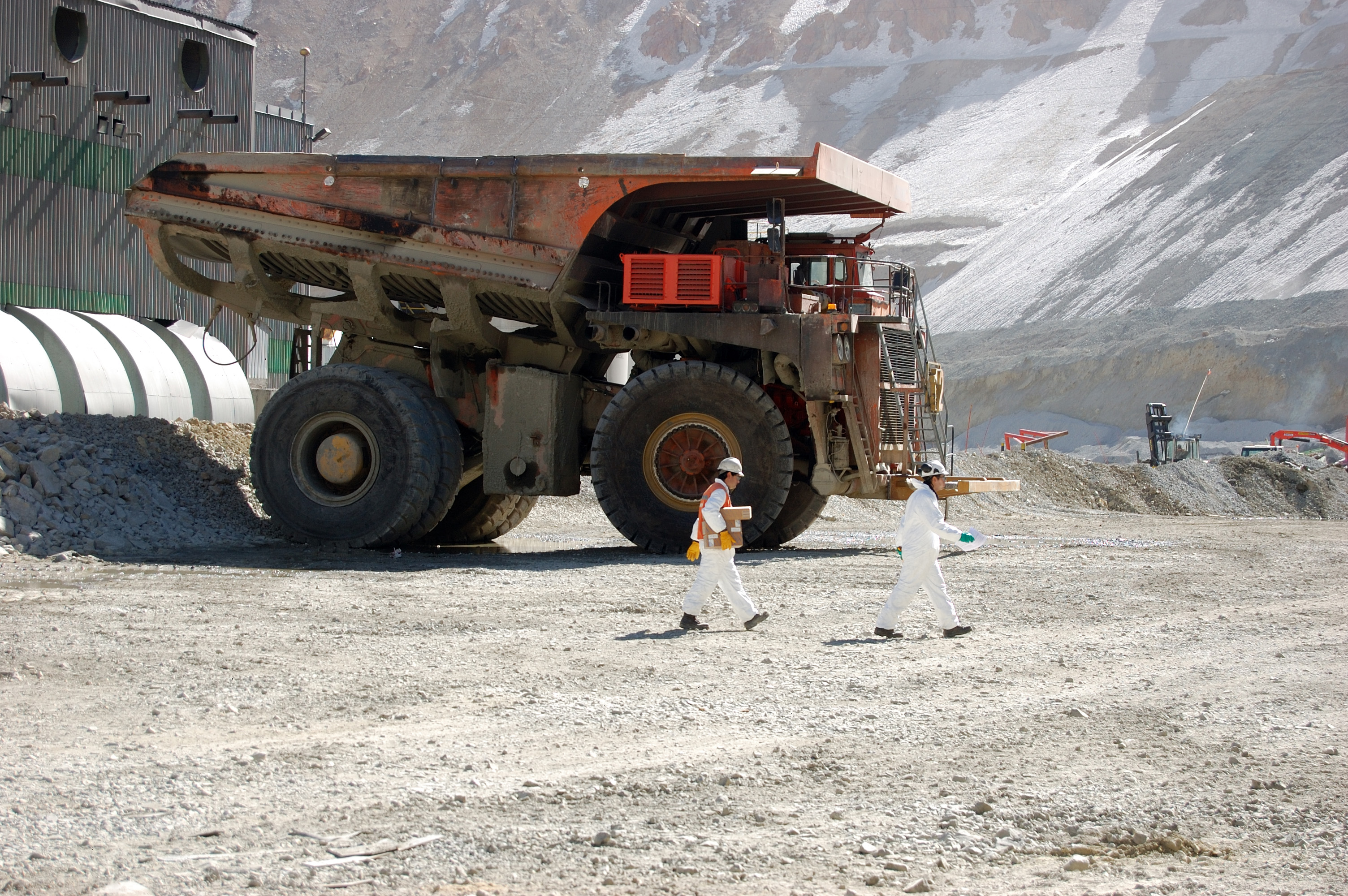 Two miners wearing white hard hats walk past a large truck at copper mine in Chile.