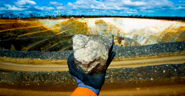 Banner: Gloved hand holds large nickel sulfide ore rock in front of an open mining pit.