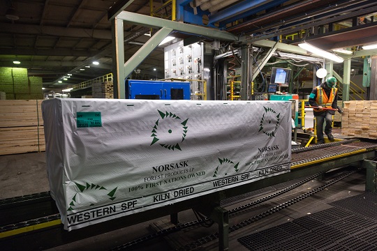 A pallet of lumber from Norsask Forest Products sits on a conveyer belt, with a worker in the background. 