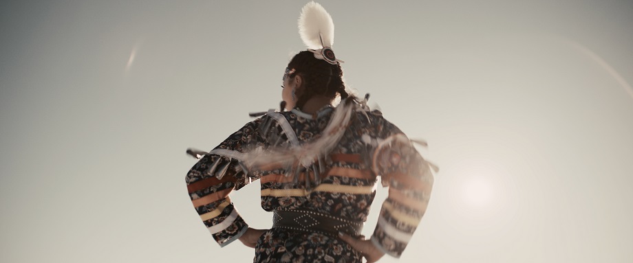 An Indigenous woman wearing a jingle dress and feathers in her hair dances outdoors, with her back to the camera.