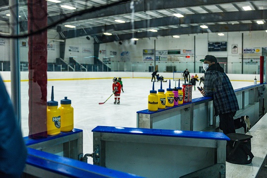 A man in a blue plaid shirt and mask stands on the bench in an arena, watching young hockey players on the ice.