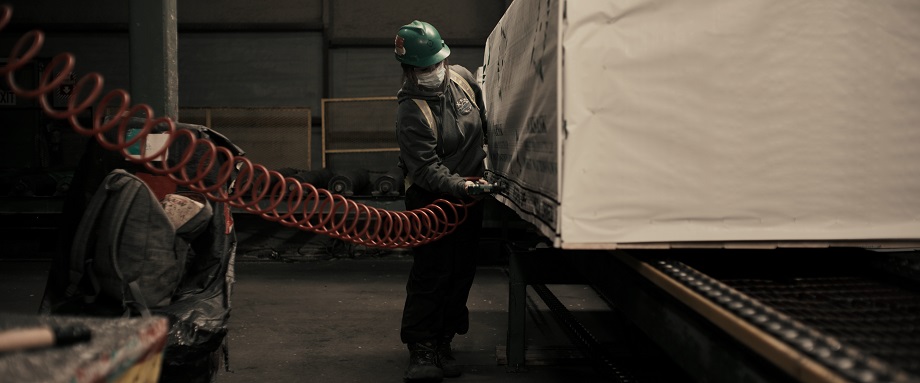 A woman in a green hardhat and grey sweatshirt uses a compression tool with a red power cord to attach the paper cover to a shipping pallet of lumber.