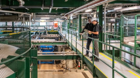Deux techniciens examinent une planche à pince devant un équipement d’Iogen conçu pour les solutions énergétiques carbonégatives.