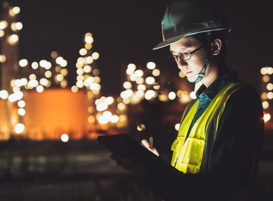 Industrial worker wearing a hard hat and high-visibility vest uses a tablet at a nighttime facility with illuminated equipment in the background.