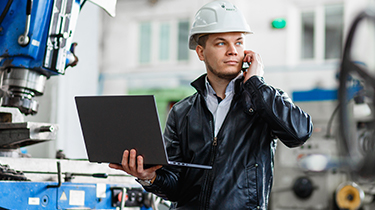 Un ingénieur, debout devant une machine industrielle avec son ordinateur portable, parle au téléphone.