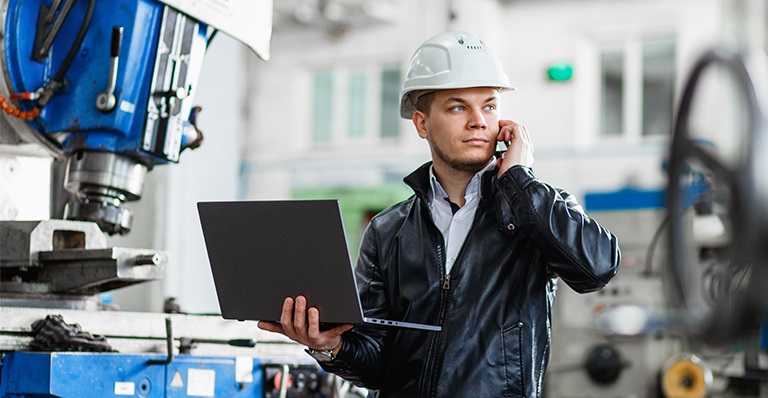 Un ingénieur, debout devant une machine industrielle avec son ordinateur portable, parle au téléphone.