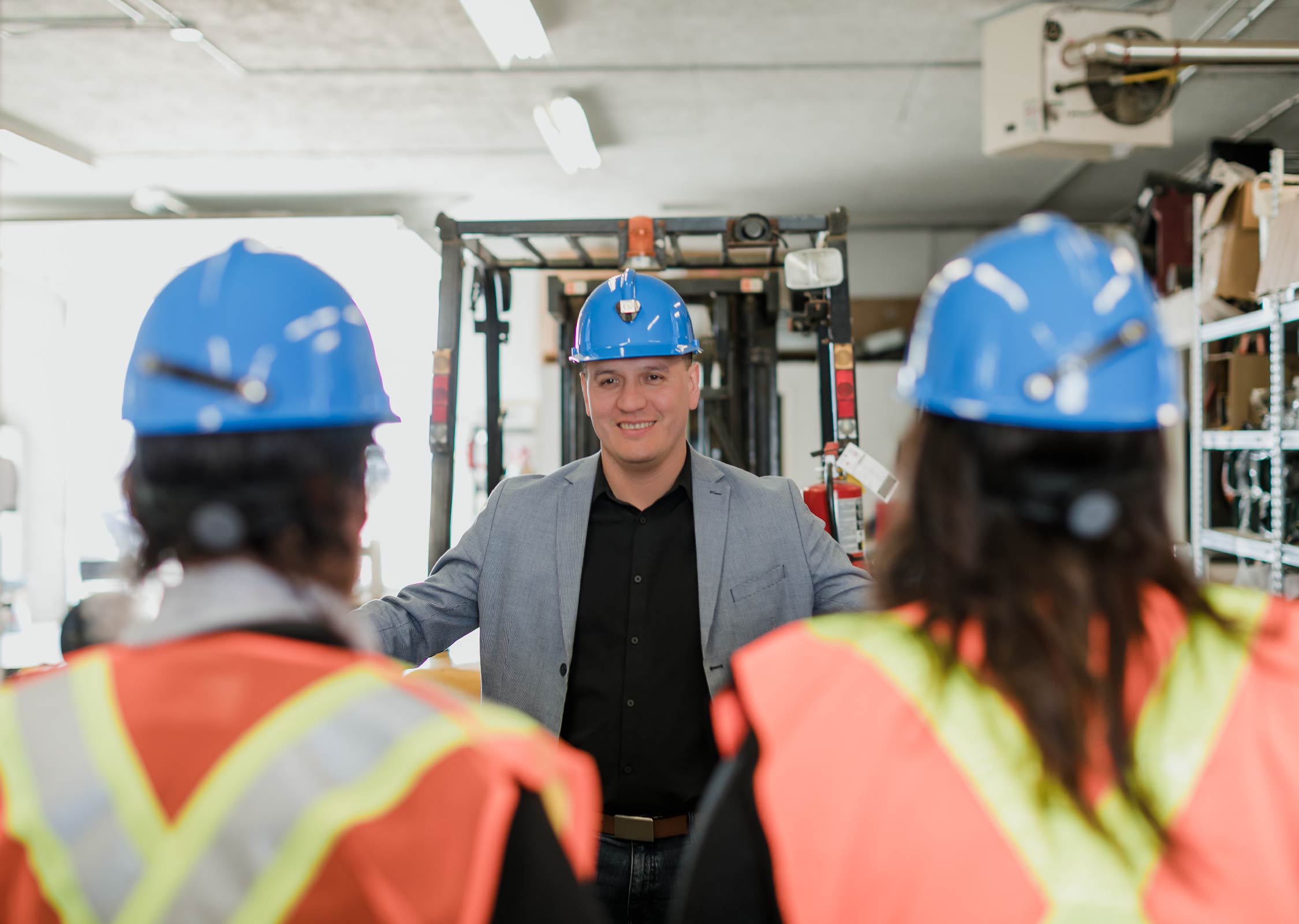 A man wearing a blue hard hat talks to two workers in a warehouse