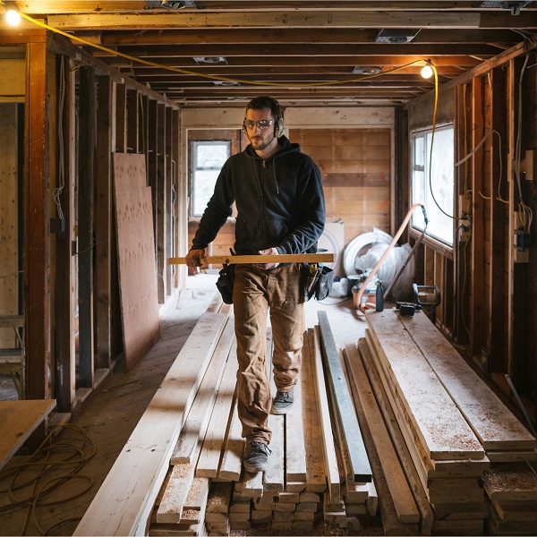 A construction worker holds a plank of wood while standing in a house under construction.
