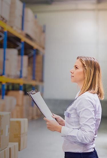 A warehouse employee taking inventory while standing in an aisle with cardboard boxes on either side.