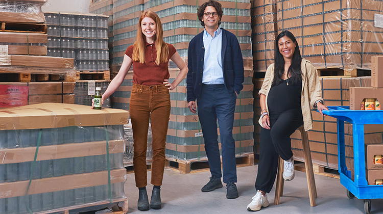 Three co-founders of an organic juice company standing in their production facility surrounded by pallets of their products.