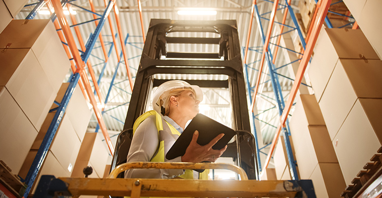 Female worker wearing hard hat on aerial platform checking inventory in warehouse.