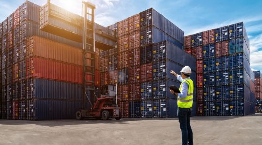 A man in a helmet and safety vest holds a clipboard and points at a forklift lifting a cargo container with his pen, surrounded by stacked containers.