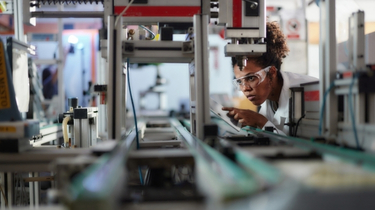 Person wearing safety glasses inspects automated manufacturing equipment on a production line, using a tablet while examining machinery components. 