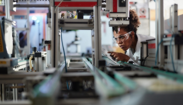 Person wearing safety glasses inspects automated manufacturing equipment on a production line, using a tablet while examining machinery components. 