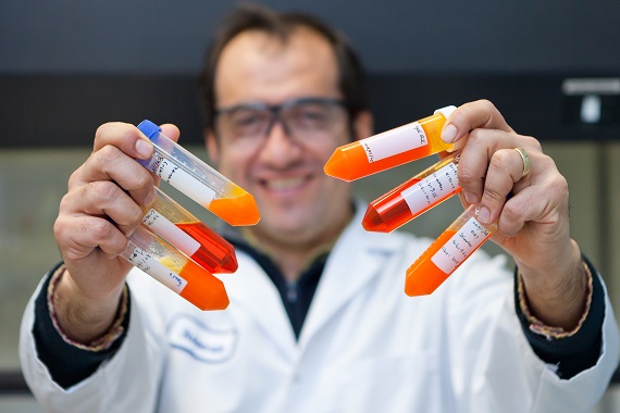 Man in white lab coat standing and holding out six test tubes containing plant-based oil.